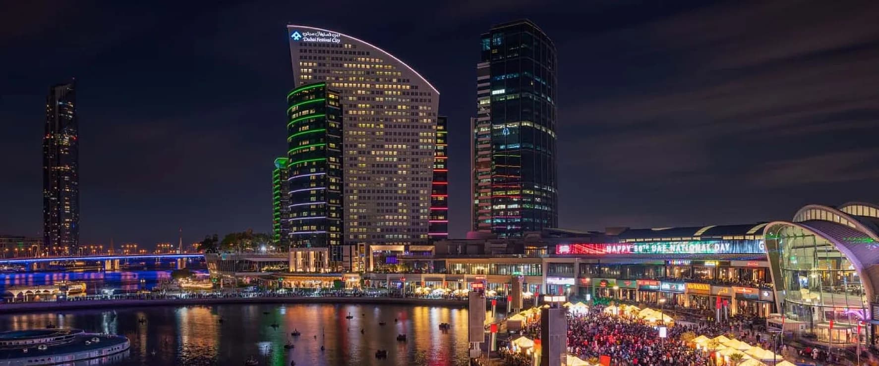 Panoramic night scene of Dubai Festival City with colourfully lit hotel towers, the mall, and a busy waterfront promenade reflected in the creek.