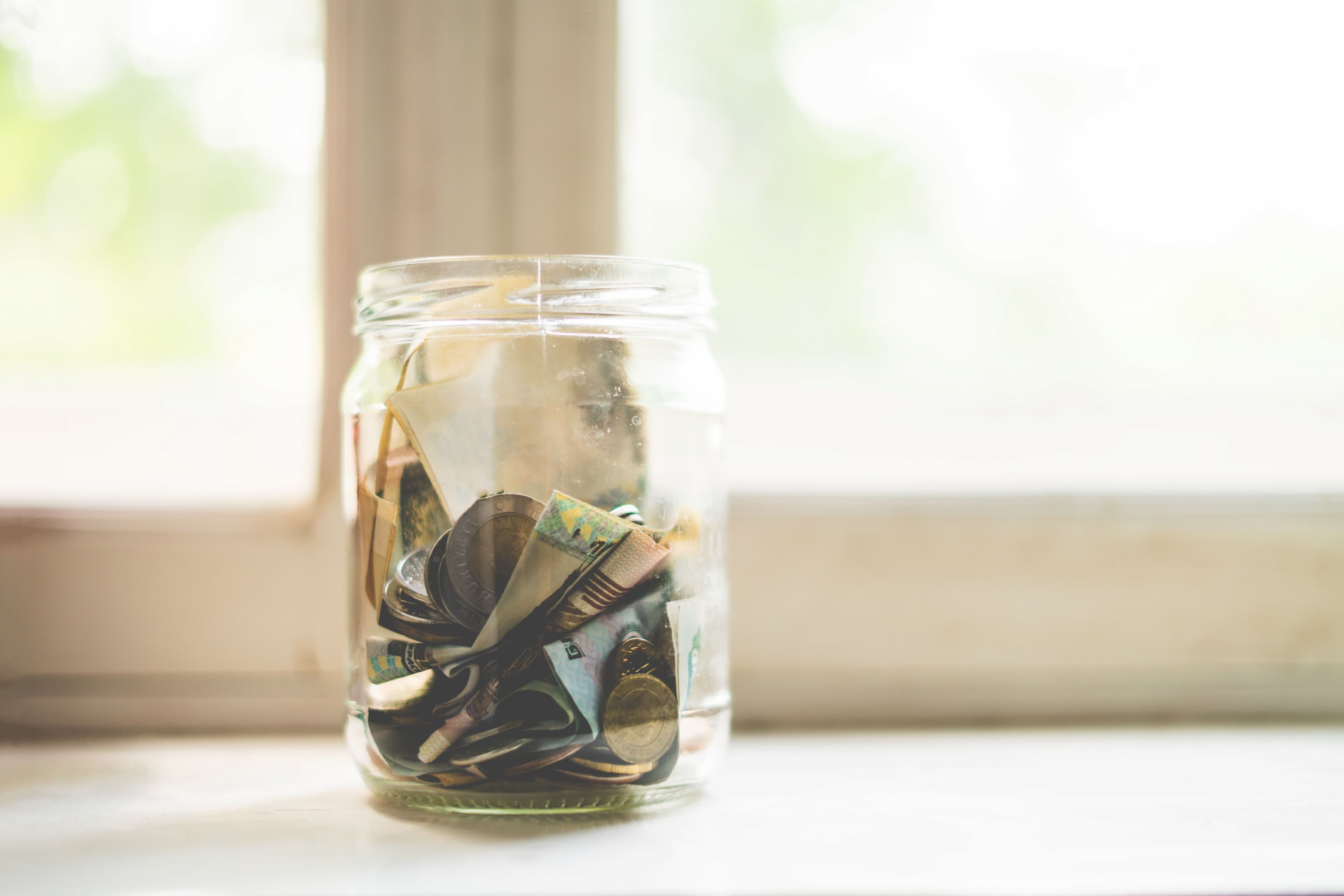 Close-up of a transparent glass jar containing assorted coins and folded paper currency near a window.