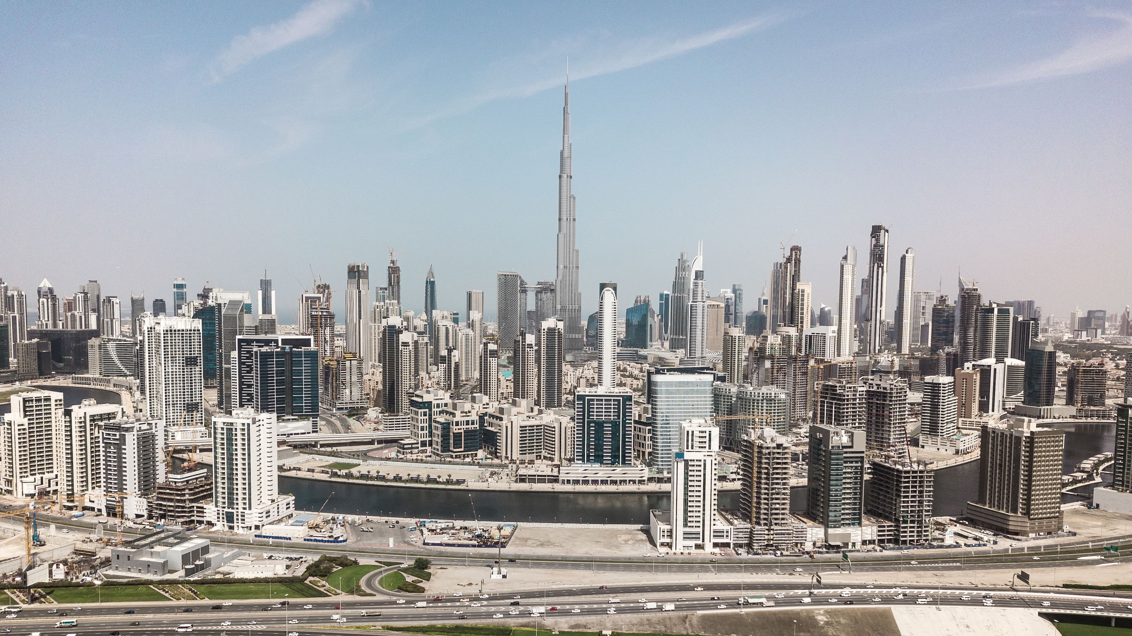 Wide-angle aerial photograph of Dubai’s downtown area, featuring the Burj Khalifa surrounded by numerous modern skyscrapers, highways, and a canal under a clear sky.